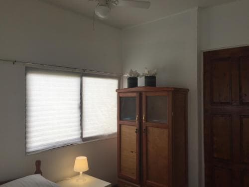 A cozy bedroom with soft lighting from a bedside lamp, featuring a wooden cabinet adorned with decorative items near a window with blinds, ideal for travelers seeking a serene and comfortable stay.