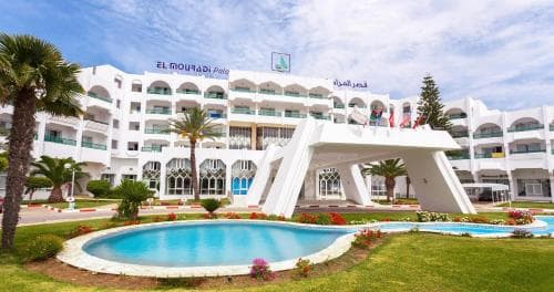 A serene tropical resort features a vibrant blue pool surrounded by lush greenery and palm trees, with a modern white building in the background, ideal for a relaxing vacation getaway.