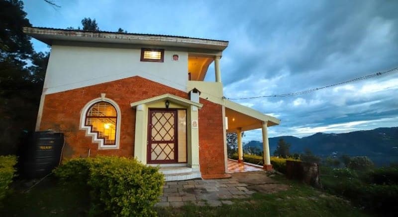 A cozy, warmly lit home with orange brick walls and a welcoming porch sits nestled among lush greenery against a dramatic sky, ideal for travelers seeking a serene retreat in Tamil Nadu's scenic landscapes.