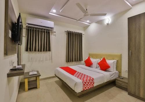 A cozy hotel room features a neatly made bed with red accents, wooden furniture, and modern amenities like a mounted TV and air conditioning, ideal for travelers seeking comfort and convenience.