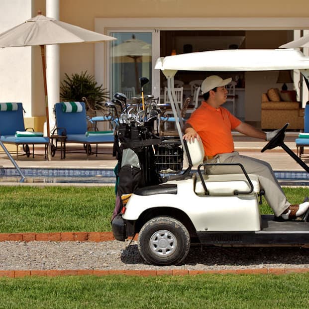 A golfer drives a cart loaded with clubs past a serene resort poolside area, ideal for leisure travelers seeking relaxation and recreation.