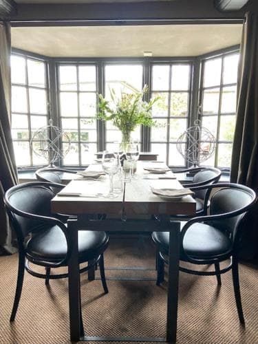 A cozy dining area with a wooden table set for four, surrounded by black chairs and large windows offering a serene view, perfect for intimate gatherings or romantic dinners in a boutique hotel setting.