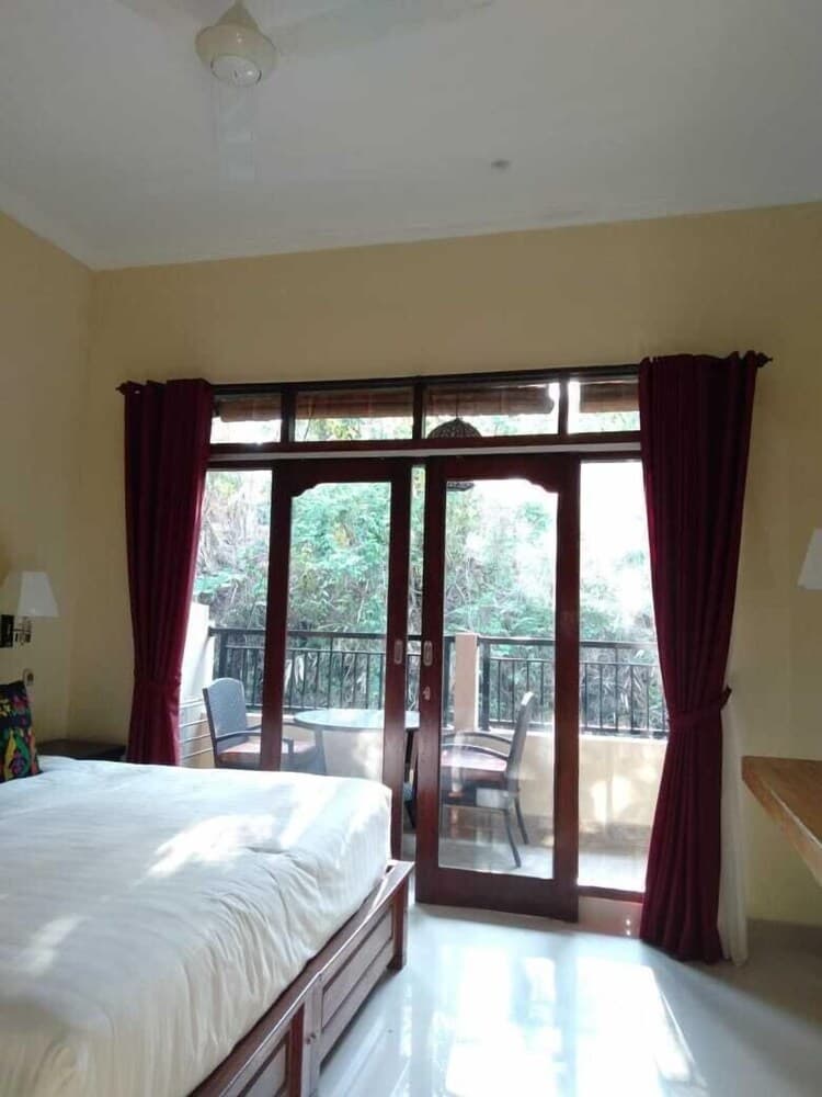 A cozy bedroom with a neatly made bed, red curtains framing glass doors leading to a balcony overlooking lush greenery, ideal for travelers seeking a serene retreat.