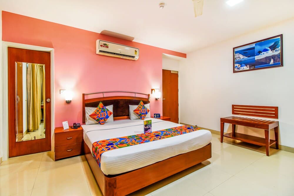 A cozy hotel room with a vibrant pink accent wall, neatly made bed adorned with colorful pillows and throw, wooden furniture, and a serene artwork creates an inviting atmosphere ideal for travelers seeking comfort and relaxation.