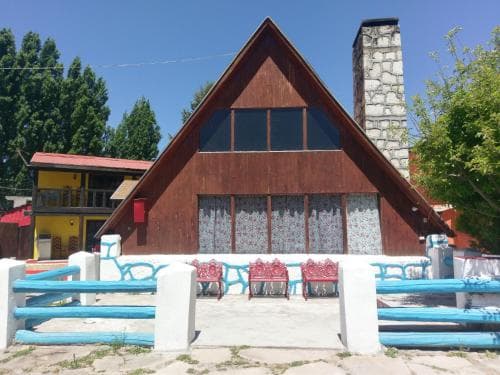A cozy wooden cabin with a stone chimney stands amidst lush greenery, featuring vibrant blue railings and red benches, ideal for a serene countryside getaway.