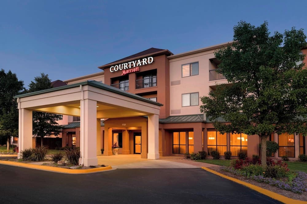 A warmly lit Courtyard Marriott hotel entrance at dusk, featuring a covered driveway, lush landscaping, and inviting windows, ideal for travelers seeking comfortable accommodations.