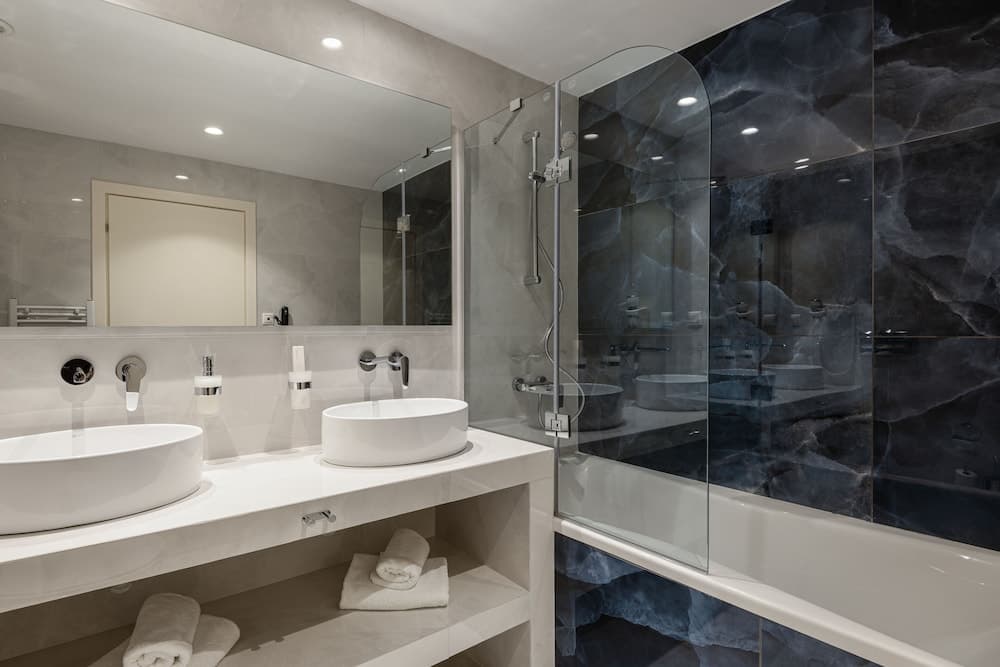 A luxurious modern bathroom features sleek white sinks, a glass-enclosed shower with dark marble walls, neatly folded towels on open shelves, and a minimalist design ideal for high-end hotel stays or boutique accommodations.