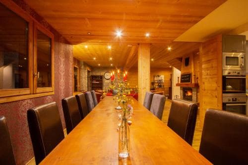 A cozy wooden dining area with a long table set for a gathering, featuring warm lighting, a fireplace, and modern appliances, ideal for a rustic retreat or family vacation.