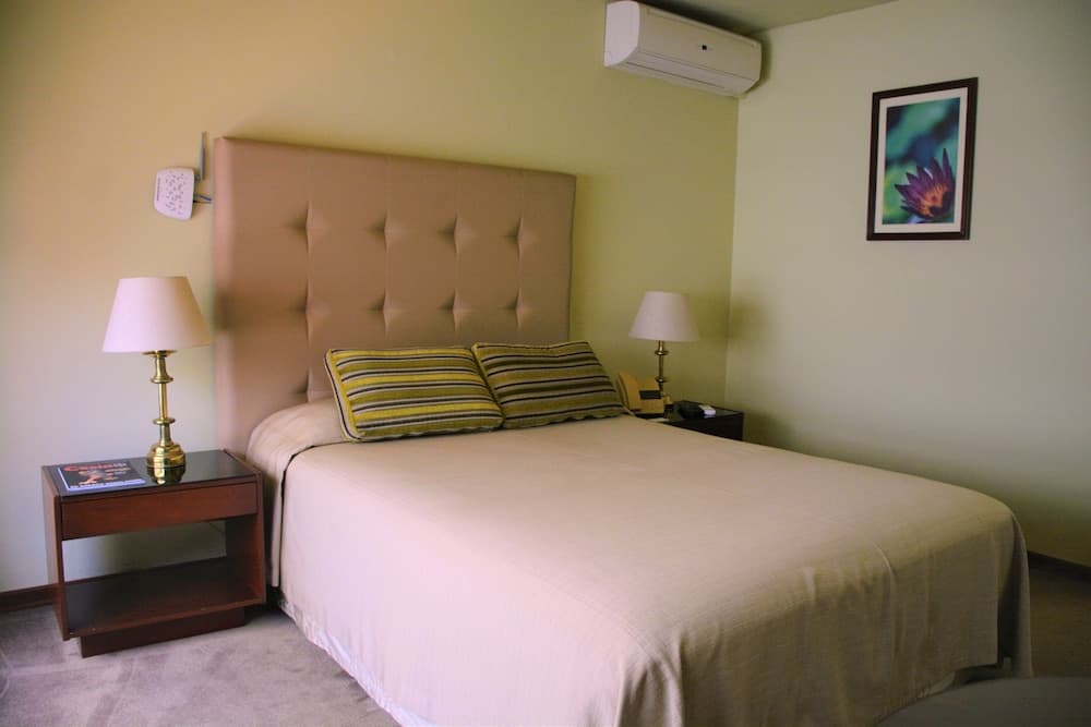 A cozy hotel room features a neatly made bed with striped pillows, wooden bedside tables with lamps, and a calming green wall, ideal for travelers seeking comfort and relaxation.