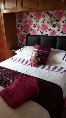 A cozy bedroom with floral wallpaper, a neatly made bed adorned with a plush toy and striped bedding, and a warm towel on the side, ideal for travelers seeking comfort in a quaint accommodation.