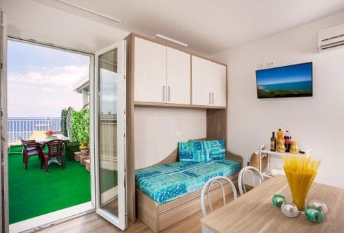 A cozy apartment with a vibrant balcony overlooking the sea offers a serene retreat, featuring modern furnishings, a colorful bedspread, and a dining setup perfect for travelers seeking comfort and relaxation by the coast.
