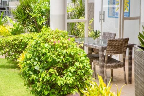 A serene outdoor patio features wicker furniture surrounded by lush greenery and vibrant flowers, ideal for relaxing or hosting small gatherings in a tropical resort setting.