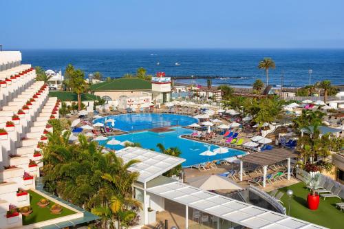 A vibrant beachfront resort features a bustling pool area surrounded by palm trees, sun loungers, and colorful umbrellas under a clear blue sky, ideal for a relaxing vacation getaway.