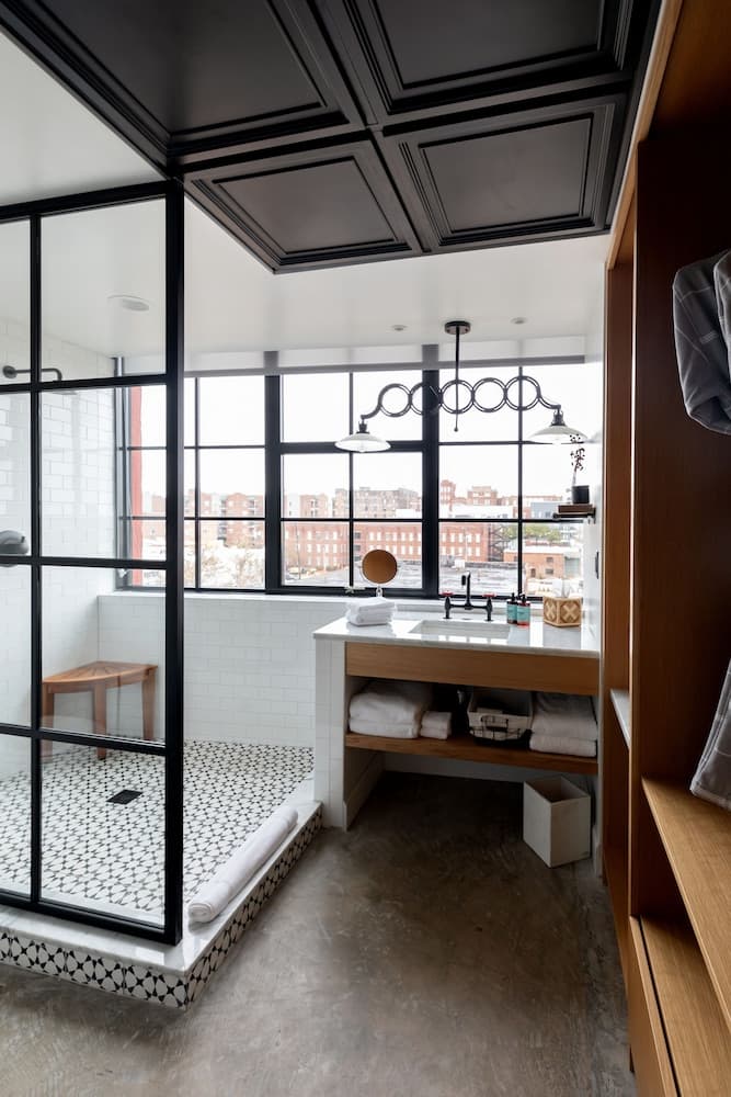 A modern urban bathroom features a sleek black-framed shower, white tiled walls, a wooden vanity with neatly folded towels, and large windows offering a cityscape view, ideal for travelers seeking stylish accommodations with a contemporary design aesthetic.