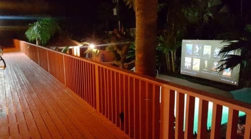 A cozy outdoor deck at night features warm lighting, lush tropical plants, and a large screen displaying images, creating an inviting atmosphere perfect for a relaxing evening gathering or event.