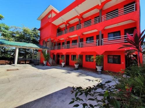 A vibrant red multi-story building with balconies stands under a clear blue sky, surrounded by lush greenery and potted plants, creating an inviting atmosphere ideal for travelers seeking accommodation in a tropical destination.