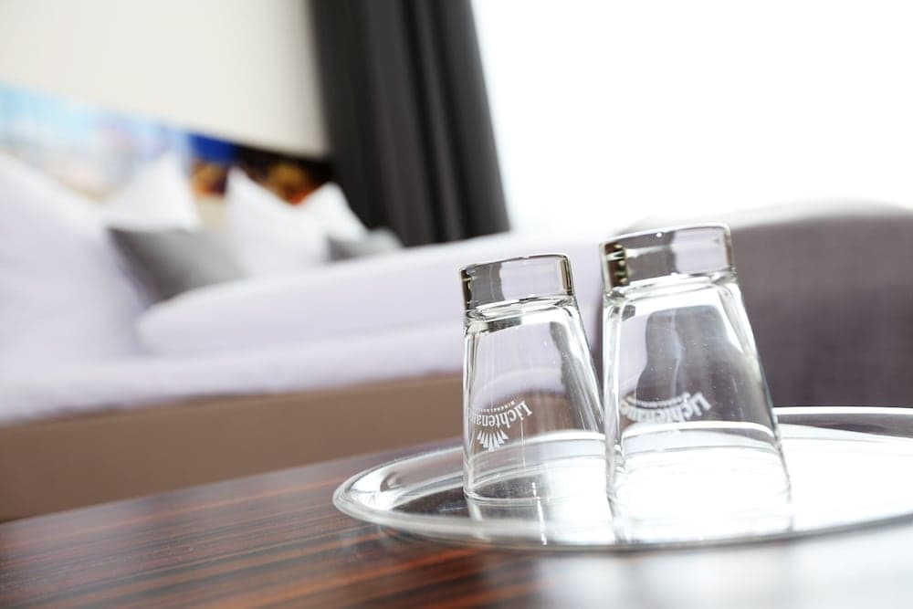 A cozy hotel room ambiance is captured with two branded glasses on a tray in the foreground, set against a blurred background of plush bedding and soft lighting, ideal for travelers seeking comfort and relaxation.