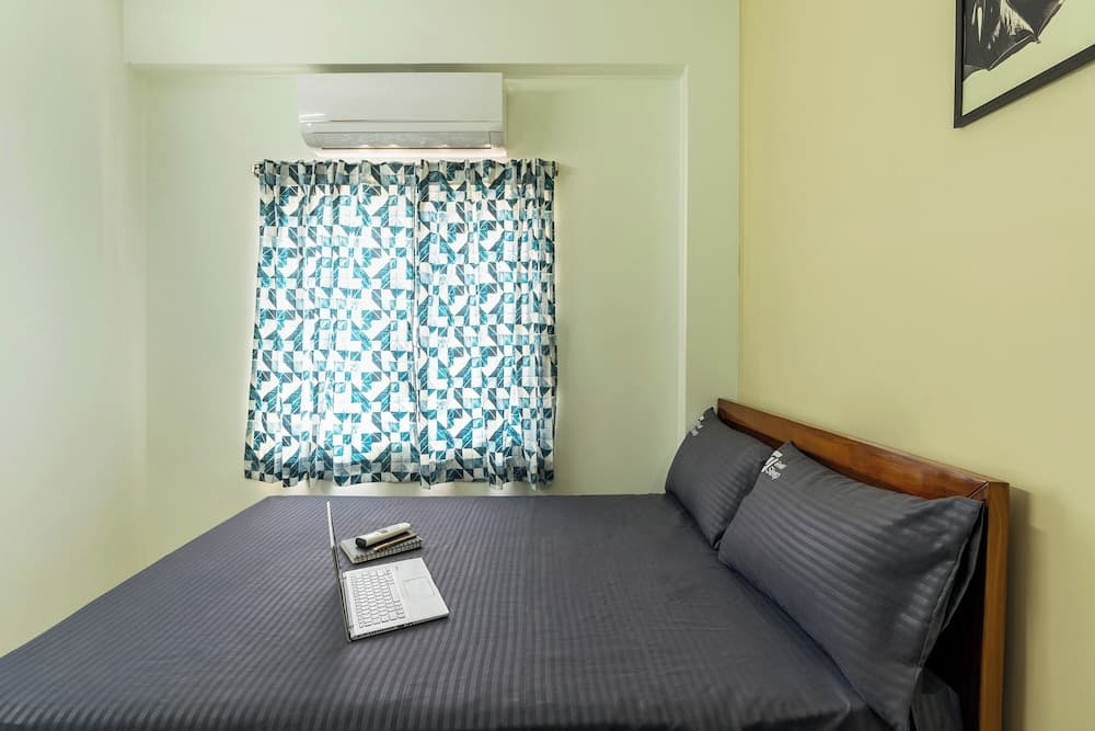 A cozy hotel room features a neatly made bed with gray linens, a laptop setup for work or leisure, patterned curtains over the window, and an air conditioner, ideal for travelers seeking comfort and functionality.