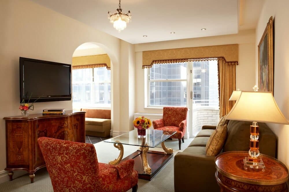 A cozy hotel suite features elegant furnishings like red patterned chairs, a glass coffee table, and a chandelier, creating an inviting atmosphere ideal for travelers seeking comfort and luxury in a cityscape backdrop.