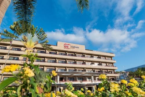 A vibrant hotel facade surrounded by lush greenery and blooming yellow flowers under a clear blue sky, ideal for travelers seeking a relaxing tropical getaway.