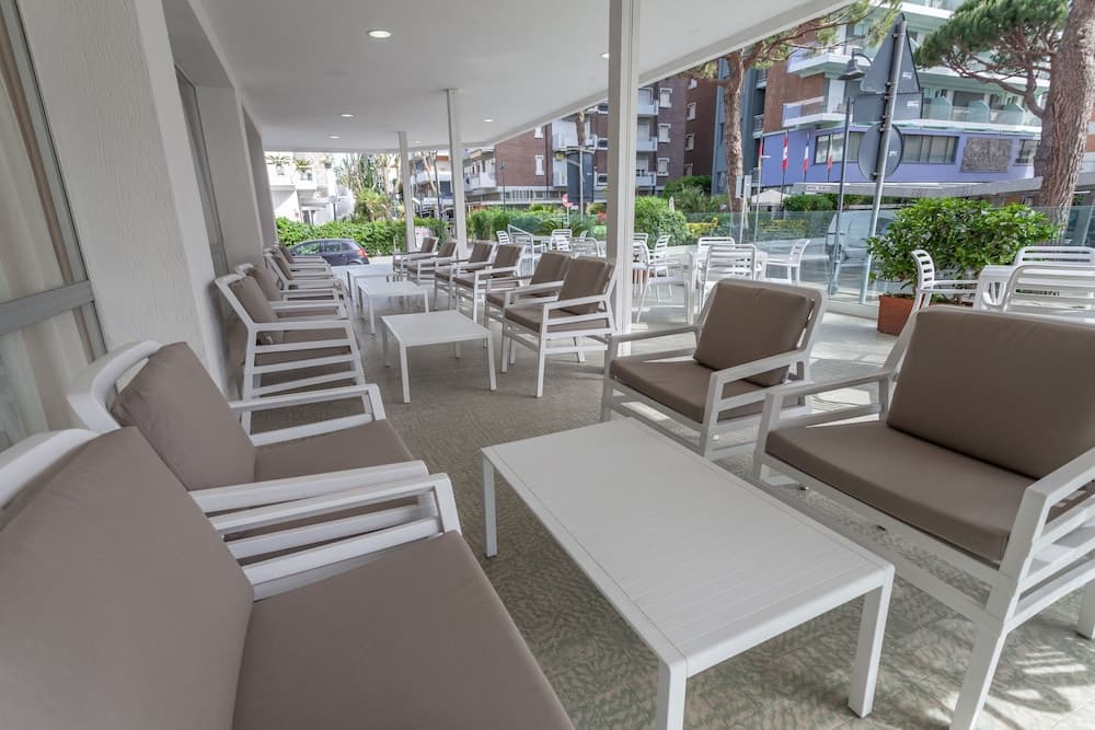 A serene outdoor lounge area with white furniture and beige cushions invites relaxation under a shaded patio, perfect for travelers seeking a tranquil retreat near urban greenery and modern buildings.