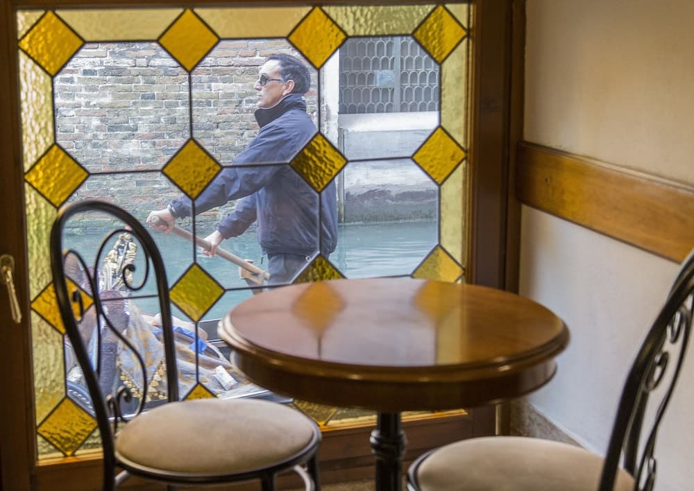A cozy café table framed by a stained-glass window overlooks a serene canal where a gondolier navigates through the picturesque setting, ideal for travelers seeking a tranquil Venetian experience.
