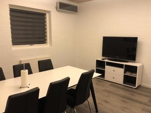 A modern meeting room with a white table, black chairs, a flat-screen TV on a white cabinet, and a window with blinds, ideal for business travelers seeking a professional space for discussions or presentations.