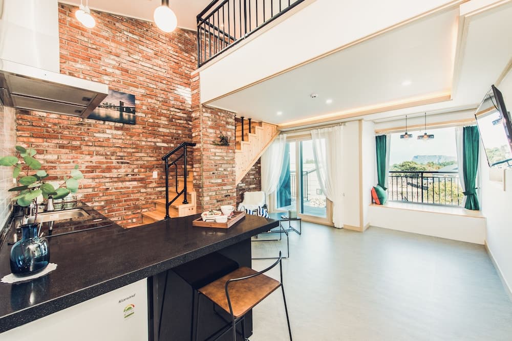 A cozy loft-style apartment features an exposed brick wall, modern kitchen bar, and a balcony with scenic views, ideal for travelers seeking a stylish and comfortable stay.