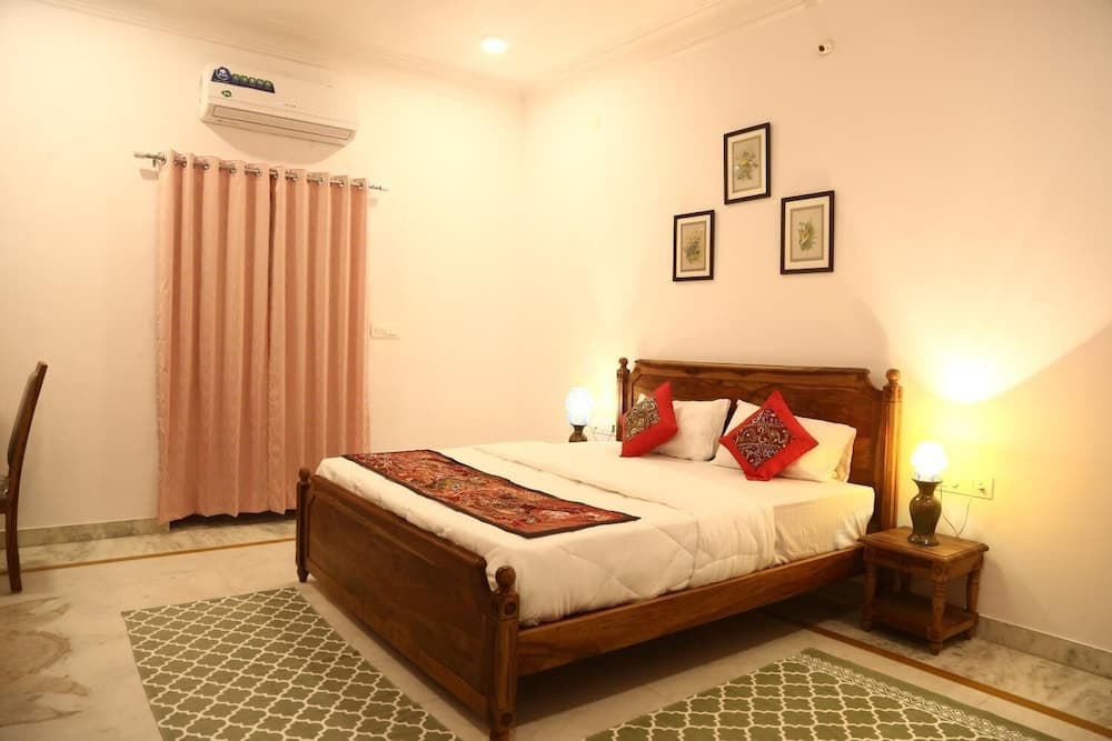 A cozy hotel room features a wooden bed adorned with red patterned pillows, warm lighting from bedside lamps, framed botanical prints on the wall, and soft peach curtains, ideal for travelers seeking comfort and relaxation.