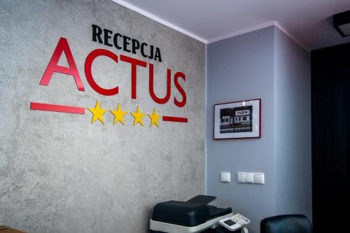 A modern hotel reception area features a bold "RECEPCJA ACTUS" sign with four stars on a textured wall, creating a professional atmosphere ideal for travelers seeking accommodation details.