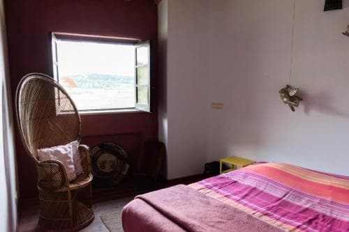 A cozy bedroom with a vibrant pink bedspread, wicker chair, and open window offering a serene view of distant hills, ideal for travelers seeking a tranquil retreat.