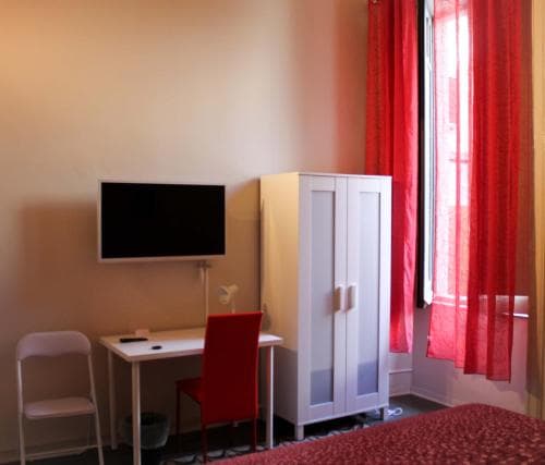 A cozy hotel room features a minimalist setup with a desk, red chair, mounted TV, white wardrobe, and vibrant red curtains by the window, ideal for travelers seeking comfort and functionality in a compact space.
