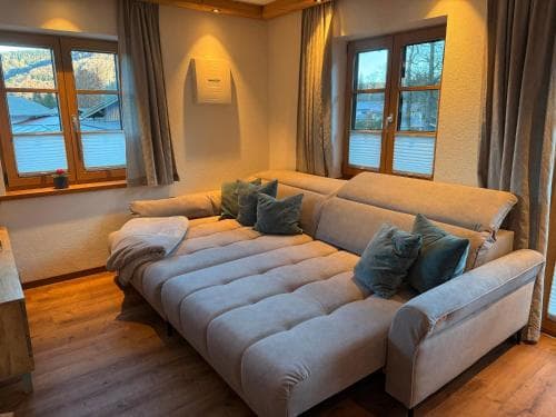 A cozy living room with a plush sectional sofa, large windows showcasing a serene mountain view, and warm wooden floors invites travelers seeking relaxation in a tranquil lodge setting.