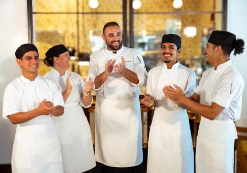 A group of chefs in white uniforms clapping joyfully in a modern kitchen setting, ideal for culinary enthusiasts seeking an engaging cooking class experience.