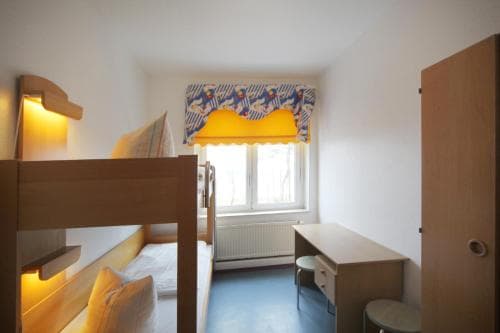 A cozy dormitory room features a bunk bed with warm lighting, a desk by the window adorned with a colorful curtain, and a serene atmosphere ideal for budget travelers seeking comfort.