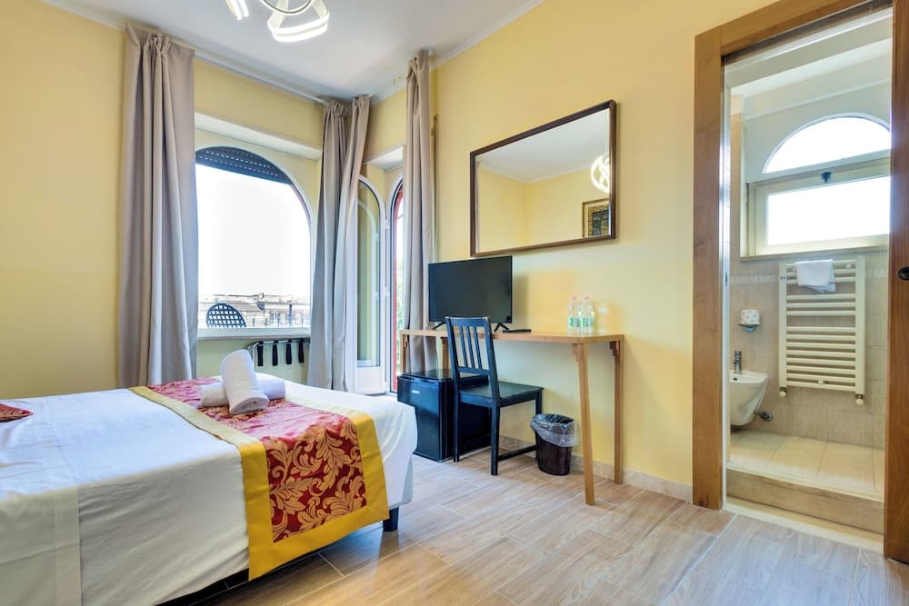 A cozy hotel room with yellow walls, arched windows overlooking a balcony, a neatly made bed with decorative linens, a desk with a TV, and an adjacent bathroom, ideal for travelers seeking comfort and convenience.