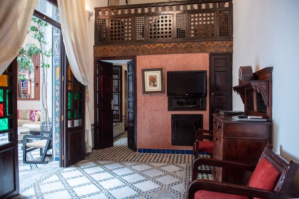 A cozy, ornately decorated room with intricate wooden lattice work, tiled floors, and vibrant furnishings invites travelers to relax in a traditional Moroccan-style guesthouse setting.