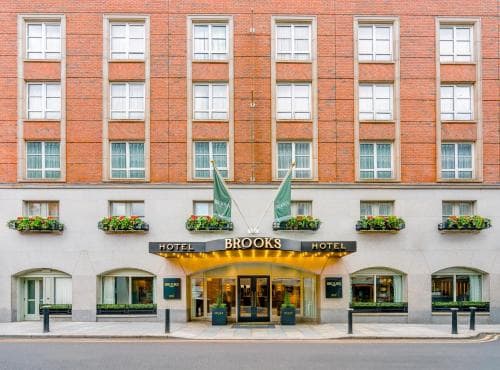 A charming urban hotel facade with brick architecture, flower-adorned windows, and a warmly lit entrance sets a welcoming tone for travelers seeking a cozy stay in a bustling city center.
