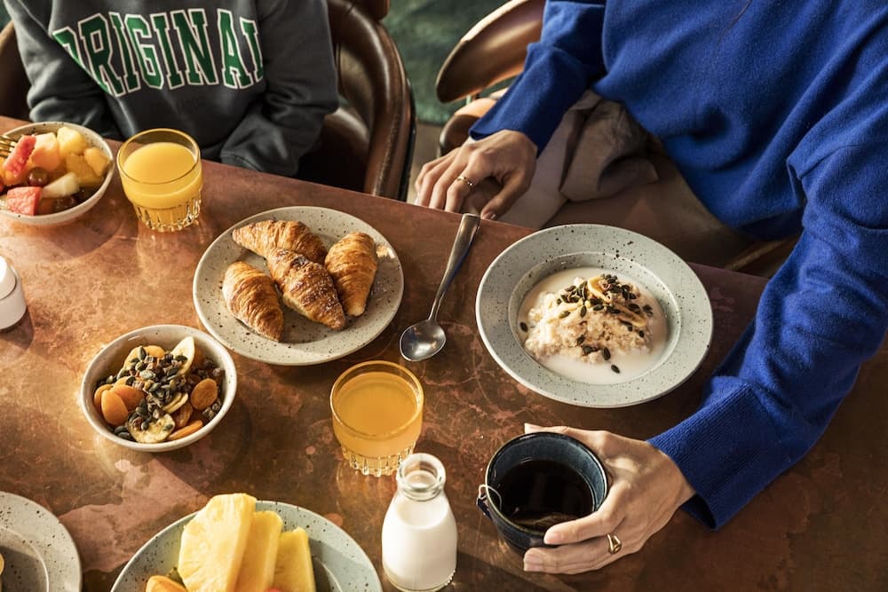 A cozy café scene features a wooden table laden with breakfast delights like croissants, fruit bowls, oatmeal, and beverages, evoking a warm and inviting atmosphere perfect for a leisurely morning outing.