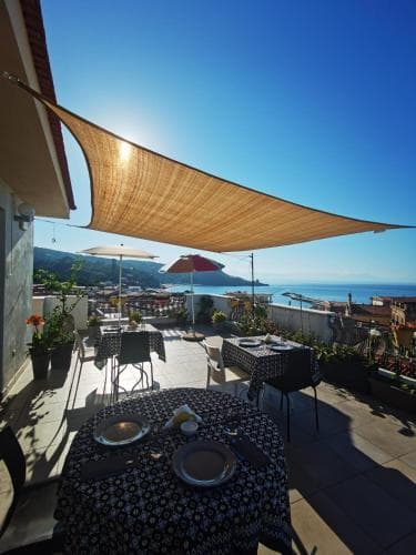 A sunlit rooftop terrace with tables set under a large shade sail overlooks the sea and hills, creating an inviting atmosphere for travelers seeking a relaxing dining spot with scenic views.