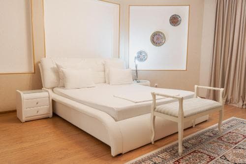 A cozy hotel room features a plush white bed adorned with pillows, a wooden chair, decorative mirrors, and a vibrant rug, creating a serene and inviting atmosphere ideal for relaxation during travel.