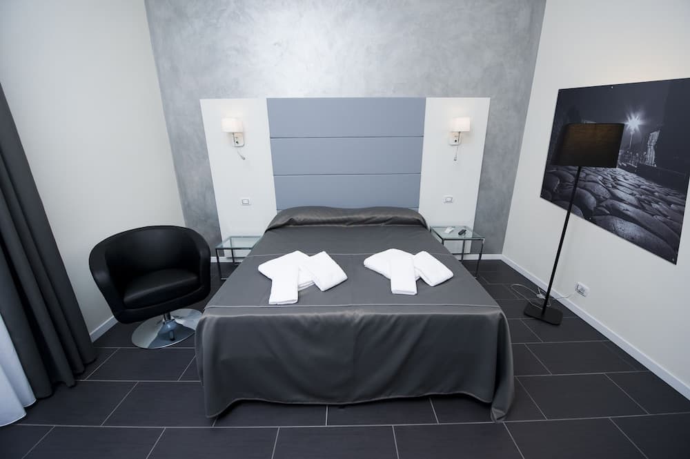 A modern hotel room features a sleek gray bed with neatly folded white towels, a black leather chair, minimalist decor, and a large wall-mounted artwork, ideal for travelers seeking a stylish and tranquil stay.