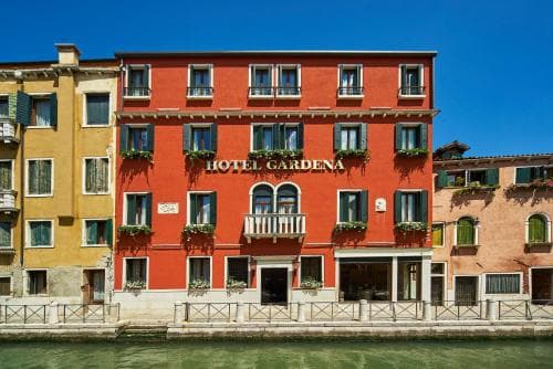 A vibrant red Hotel Gardena stands along a serene canal in Venice, featuring charming balconies and lush greenery, ideal for travelers seeking a picturesque European getaway.