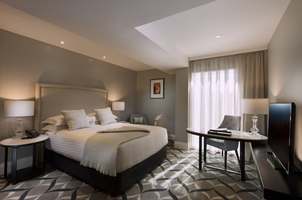A cozy hotel room with soft lighting, a neatly made bed, patterned carpet, and a desk by the window creates a serene atmosphere ideal for travelers seeking relaxation and comfort.
