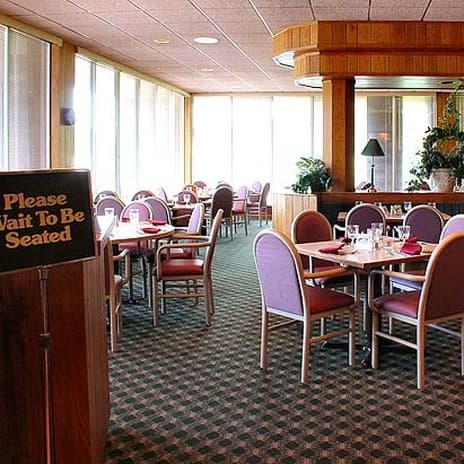 A cozy restaurant interior with ample natural light through large windows, featuring neatly arranged tables and chairs, a welcoming "Please Wait To Be Seated" sign, and a calm atmosphere ideal for travelers seeking a comfortable dining experience.