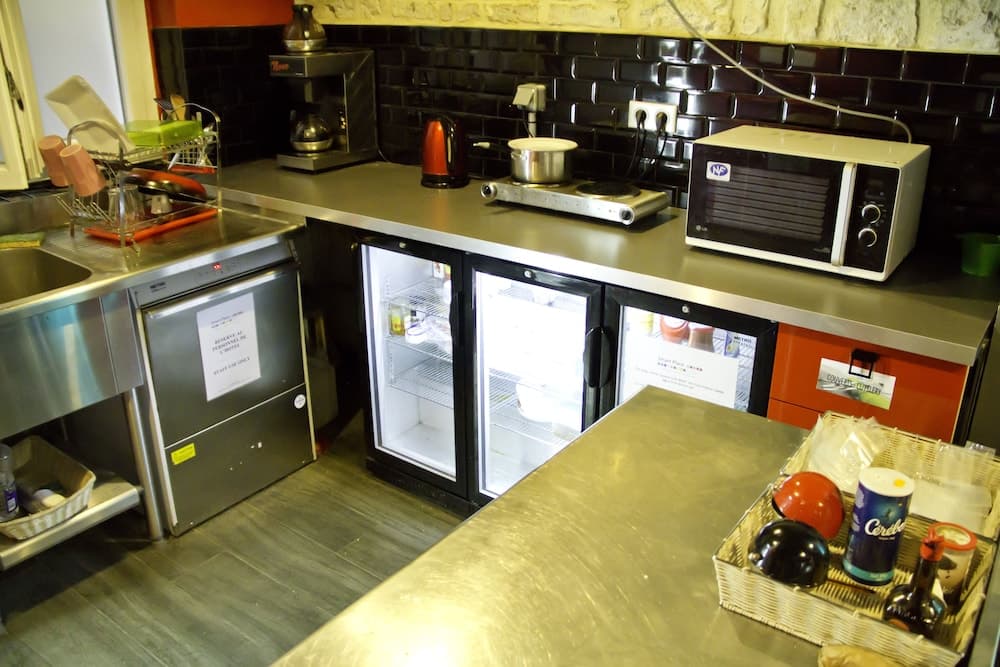A cozy communal kitchen with black tiled walls, stainless steel appliances, and a fridge stocked with essentials, ideal for travelers seeking a self-catering experience in a shared accommodation setting.