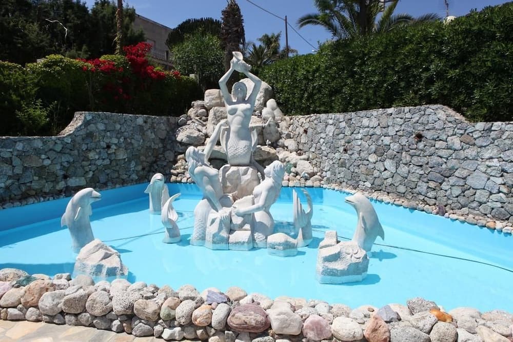 A serene garden fountain features a central mermaid statue surrounded by dolphins, set against a stone wall backdrop with lush greenery, ideal for tranquil vacation photo spots.