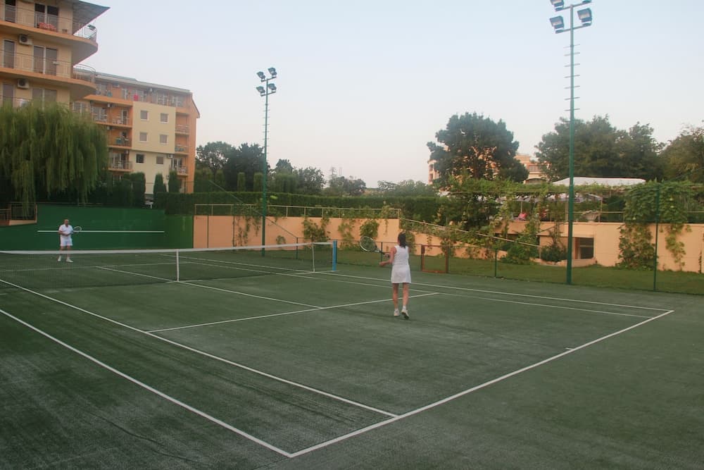 A serene outdoor tennis court surrounded by lush greenery and residential buildings offers an ideal spot for sports enthusiasts to enjoy a game under the soft glow of evening lights.