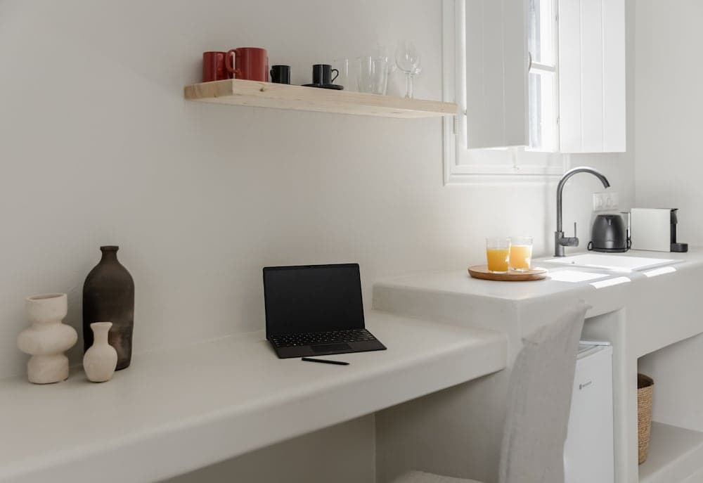 A serene minimalist workspace features a sleek white desk with a laptop, decorative vases, and a cozy kitchenette with orange juice glasses, ideal for travelers seeking a calm and functional retreat.