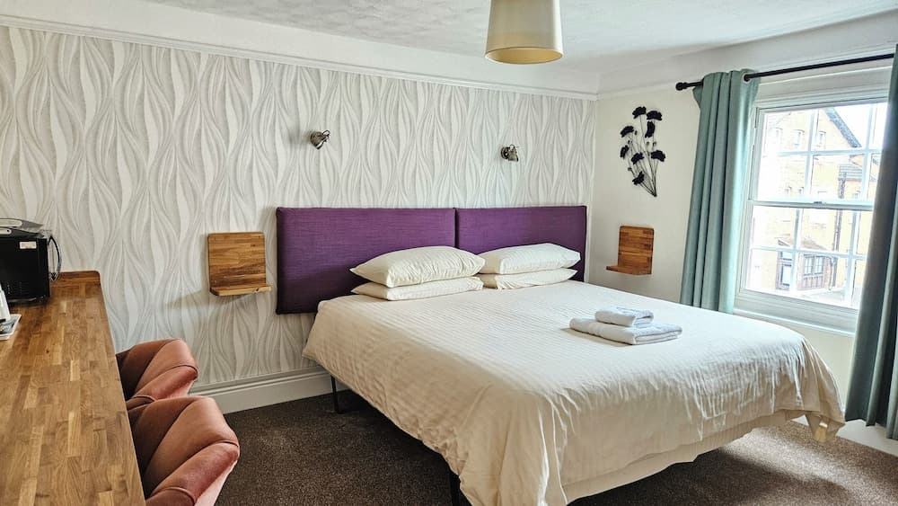A cozy hotel room features a neatly made bed with purple headboards, soft lighting, wooden accents, and a window view of urban buildings, ideal for travelers seeking comfort and relaxation.